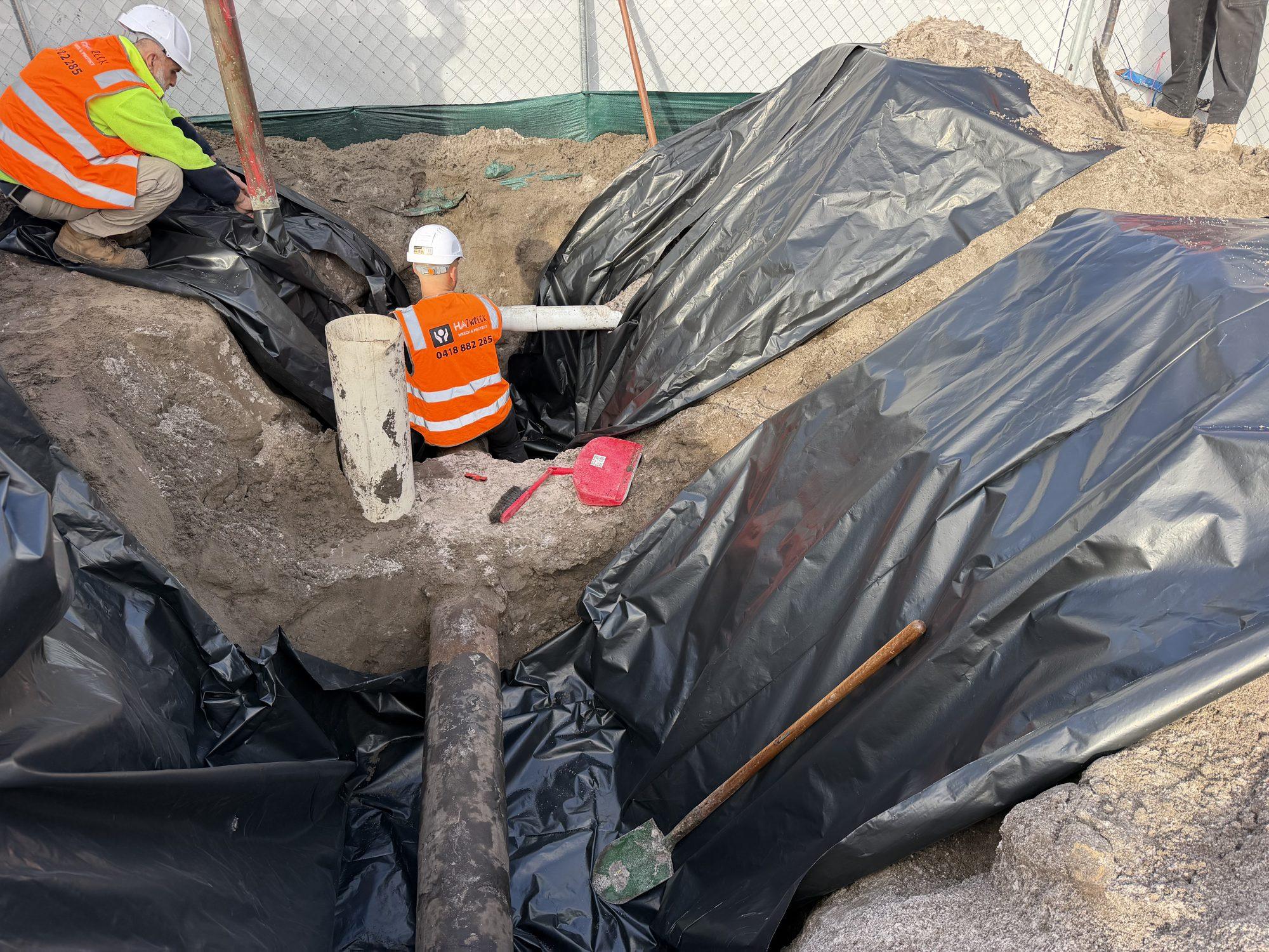 Crew member working over the lined excavation with heavy plastic sheeting around the pipe