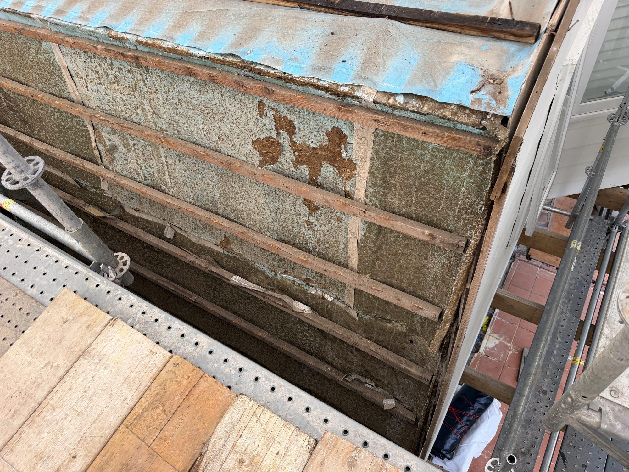 Original fibro wall cladding with peeling paint layers exposed mid-removal behind scaffold