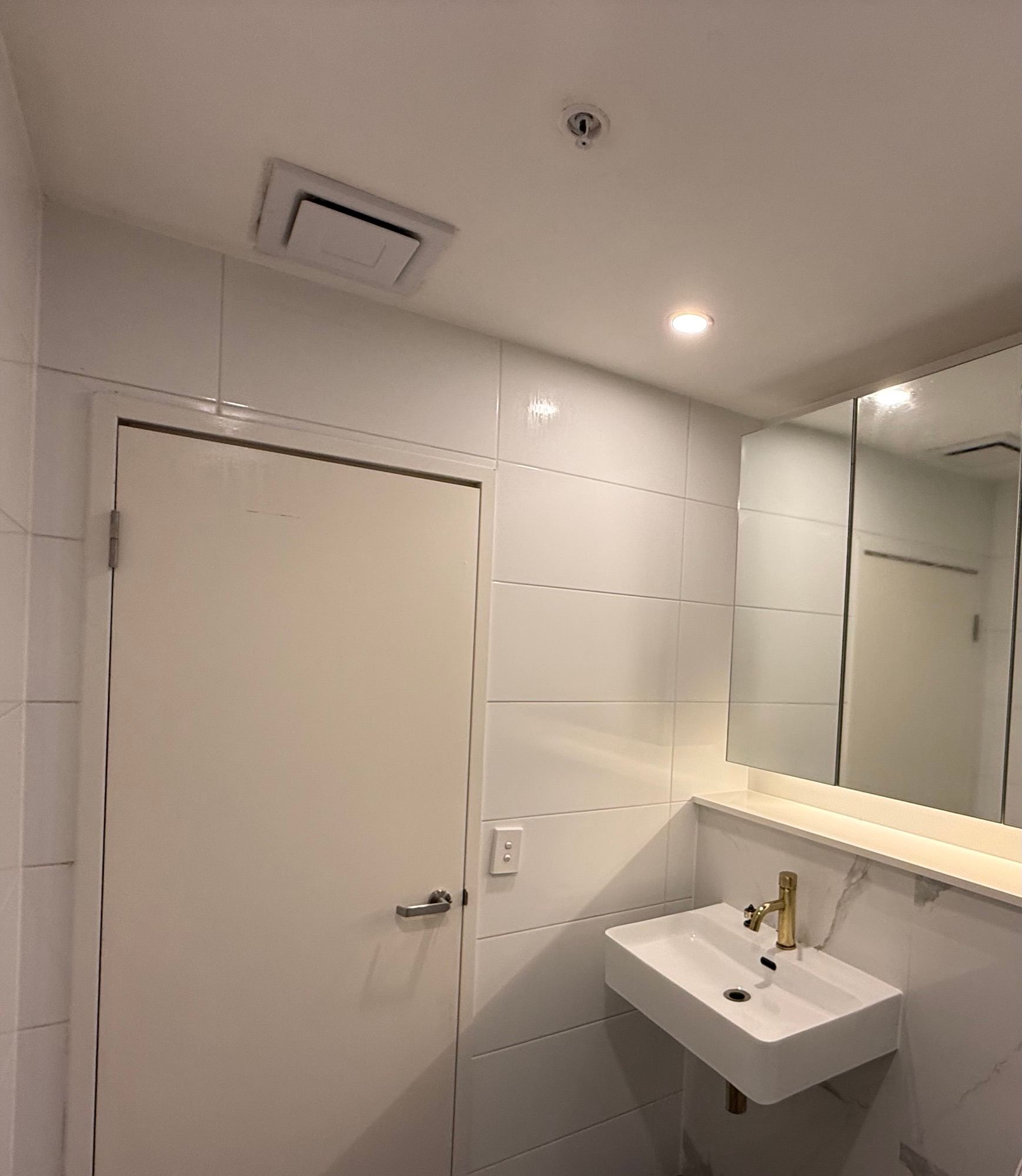 The same bathroom from the same spot after remediation: clean white ceiling, exhaust fan, door, mirror cabinet and basin