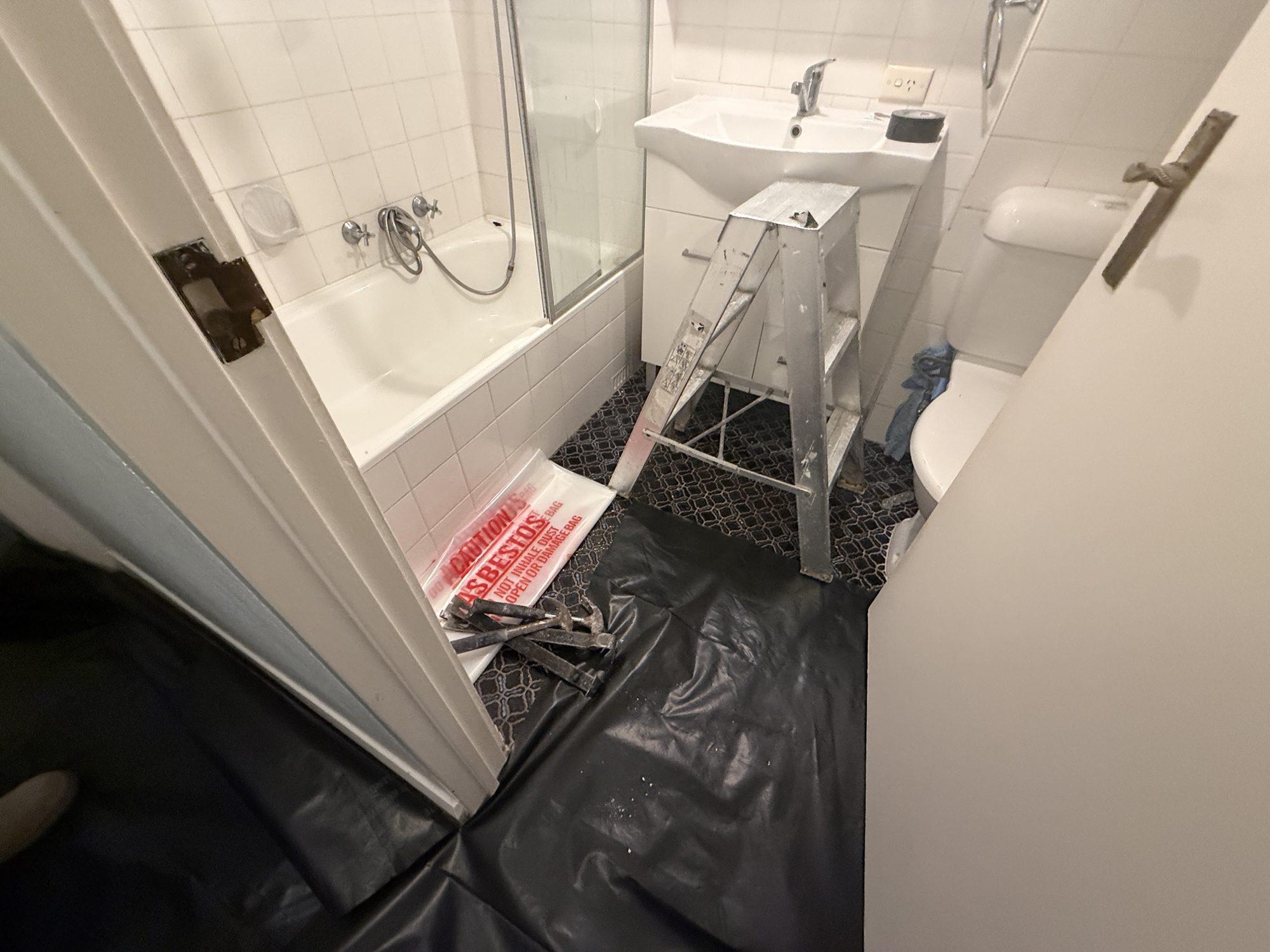 Bathroom floor under plastic with an asbestos warning sheet and a step ladder before panel removal